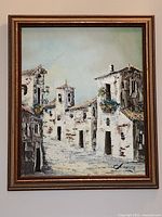 Framed oil painting showing village street scene with buildings, flowers, and signature at bottom right.