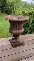 Brown plastic urn-shaped outdoor planter on wooden table, set against garden background