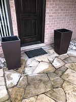 Two brown resin tall square planters on a stone patio in front of a door.