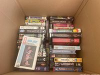 View of multiple VHS tapes inside a box showing movie titles including Career Opportunities, Sixteen Candles, Winnie the Pooh, The Musketeers, Two Moon Junction, and others.