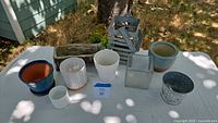 All nine pots and containers arranged on a white tablecloth, displaying variety of styles including ceramic, glass, and metal items suitable for planting and decorative use.