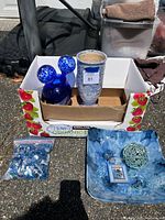 Lot contents overview showing blue ceramic vase, blue glass decorative piece, bag of decorative beads, floral fabric with woven ball and small boxed item.