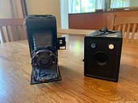 Two antique cameras on wooden table: Kodak folding camera with bellows open and Ansco box camera closed side by side.