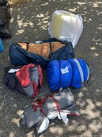 Four sleeping bags in blue, red, black, and gray colors, along with white foam sleeping pads lying on the ground outside in partial shade.