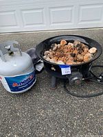 Photo of propane tank and firepit with rocks on pavement in front of garage door, showing tank label and firepit hose connection.