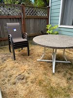 Four brown fabric mesh stackable outdoor chairs next to a round outdoor table with metal legs on dry grass next to wooden fence and house siding.