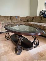 Side perspective of the oval coffee table showing glass top, wood frame with scrollwork legs and lower wooden shelf