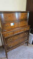 Full view of vintage chest of drawers made of burled wood with five drawers and brass handles. Shows wear and scratches on surface.