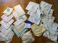 Top-down view of over 20 vintage handkerchiefs arranged on a wood floor showing a variety of white, ivory, and pastel colors with lace and embroidered details.