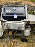 Front view of Weber Spirit II propane grill showing black lid with thermometer, two control knobs, side shelves, propane tank, and wheels.