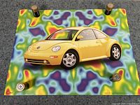 Top view of the yellow VW New Beetle poster held down by four batteries, showing the bright car with a psychedelic colorful background.