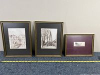 Three framed sketched artworks side by side on a blue carpet, showing approximate size with a measuring tape. Frames are gold-toned. Each sketch features buildings and architectural scenes.