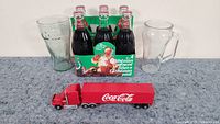 Full view of all items showing vintage 6-pack bottles in Christmas carrier, two Coca Cola branded glasses, and red Coca Cola toy truck.