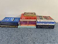 Photo of 14 cookbooks arranged in two uneven stacks on floor against wall, titles highlighting regional cuisines like Kentucky, Louisiana, Mississippi, New England, Chinese, Italian, and Mexican cooking.