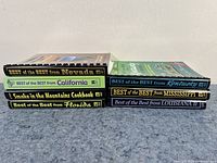 View of seven hardcover cookbooks stacking horizontally showing spine titles featuring food heritage from various U.S. states like Nevada, California, Kentucky, Mississippi, Louisiana, and Florida.