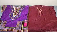 Two cultural shirts laid flat, one purple with multicolored designs and one maroon with golden motifs