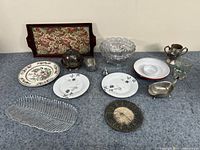 14 assorted dining room items on a dark surface against a plain wall, including plates, bowls, silver serving pieces, and a wooden framed fabric panel.