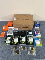 Photo of printer ink cartridges, toner cartridges in box, and typewriter supplies arranged on table.