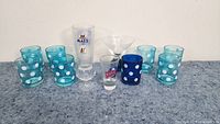Wide shot showing the full set of 12 small glasses on a grey surface against a white wall. Includes turquoise polka dot glasses, dark blue polka dot glass, Maes glass, Pepsi shot glass, martini glass, and small shot glass.