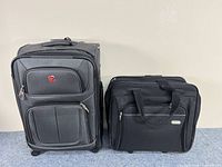 Two black travel bags side by side on a blue carpeted surface against a plain beige wall: Swiss Gear carry-on suitcase and Travis laptop bag.