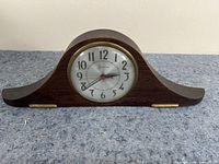 Front view of vintage wooden mantle clock with white face and Arabic numerals, black hour and minute hands and a red second hand, sitting on grey surface.