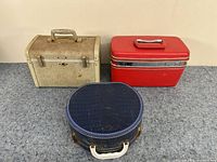 Three small vintage travel cases: beige rectangular with metal handle, red rectangular hard case with silver band, navy blue oval with white handle