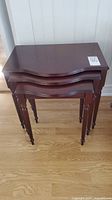 Stacked view of three wooden nesting tables showing dark cherry finish, curved fronts, and turned legs.