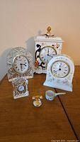 Full lot shown: 4 decorative clocks in glass and ceramic, one miniature telephone and small ceramic ladle and bowl, all arranged on wooden surface.