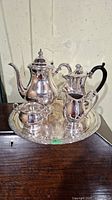 Silver plated coffee set on a wooden surface with hot water pot, coffee pot with black handle, creamer, sugar bowl, and round tray.