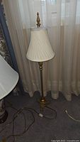Full view of the brass floor lamp showing white fabric pleated lampshade, finial, and power cord with switch lying on carpeted floor in front of curtains.