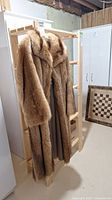 Full view of a medium to light brown fur coat hanging on a wooden rack, showing long sleeves and wide collar.