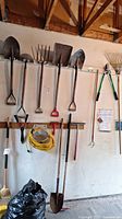 Wide view showing wood and metal shovels, pitchfork, edger, extension cord, hedge shears, and squeegees hanging on garage wall.