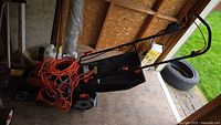 Black & Decker electric lawn mower with coiled orange extension cords on top, placed inside a wooden shed.