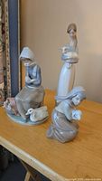 Three Lladro porcelain figurines of girls with lambs shown together on a table to illustrate lot contents and condition.