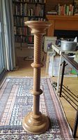 Full view of the turned wooden plant stand showing the spiral design on the stem and round base, standing on a patterned rug in a room with bookshelves and a fireplace.