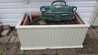 Photo showing beige rectangular planter with reddish-brown plastic liner and green metal car front garden decor inside with soil.