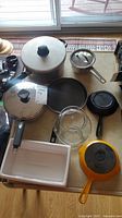 Top-down photo of all cookware and glass items arranged on a table including pans, pot, glass vase, and ceramic dish