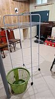 Metal clothes drying rack and green plastic laundry basket placed on the floor indoors with some furniture in the background.
