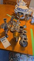 Photo of various silver plated items on a wooden table including candle holders, wine goblets, sugar shaker, condiment containers, salt and pepper shakers, cup, and tray.