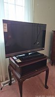 Sony flat screen TV standing on a wooden side table by a window with curtains. The screen is black and the bezel is black with a silver Sony logo on the frame.