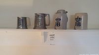 Four beer steins displayed on a white shelf including two pewter and two ceramic steins.