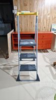 Front view of the Cosco metal step ladder with yellow top handle and three black plastic steps.