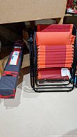 Folded red and orange folding lounger with black metal frame next to a navy blue fabric carry bag for the sun shelter.