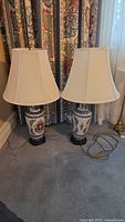 Two matching porcelain table lamps with cream-colored bell-shape fabric shades placed on carpeted floor near window with patterned curtains.