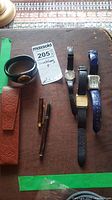 Four wristwatches and one fountain pen with matching case laid out on a wooden surface, includes lot tag