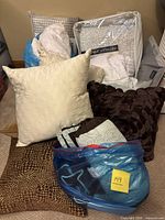 Photo showing assorted pillows in various fabrics including quilted cream, petal-like dark brown, and animal print pillows with a packaged Fabienne Vittadini duvet cover and other bedding stacked on carpet.