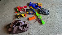 Six assorted NERF toys and toy items displayed on a concrete floor with one fabric bag and a gray glove visible