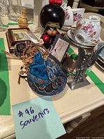 Photo showing assorted small household souvenirs and decor on a table, including a wooden Japanese doll, floral teacups, carved wooden box, fabric pouch, framed picture, and metal figurine.