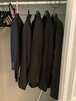 Photo showing five men's blazers hanging on a rack inside a closet, viewed from the side, several in shades of black and dark gray.