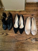 Three pairs of women's shoes on wooden floor including black ankle boots, black pointed toe heels, and beige ballet flats, all size 7.